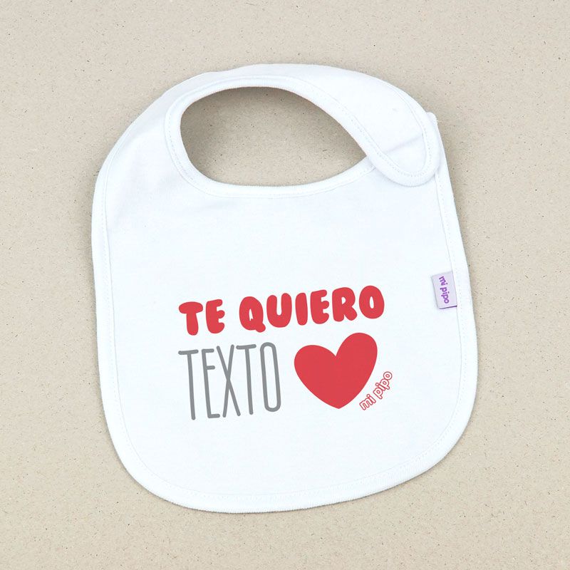 Babero blanco de tela con cuello redondo, texto "Te quiero" en rojo, "TEXTO" en gris y corazón rojo.