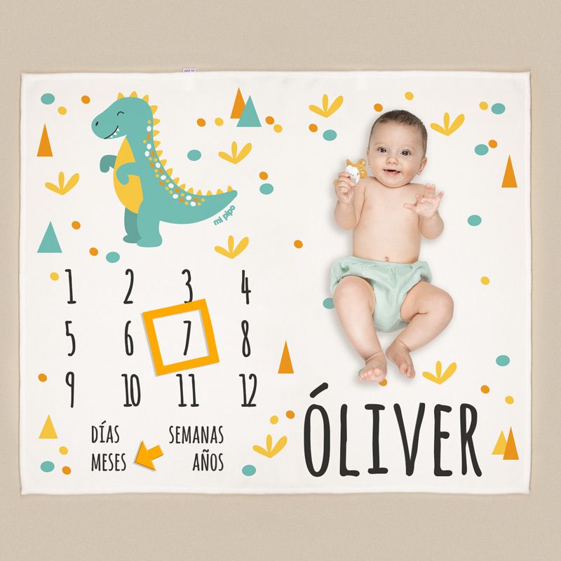 Manta blanca con dinosaurio verde y detalles naranjas, calendario para cumplemes con número resaltado y nombre Óliver en.